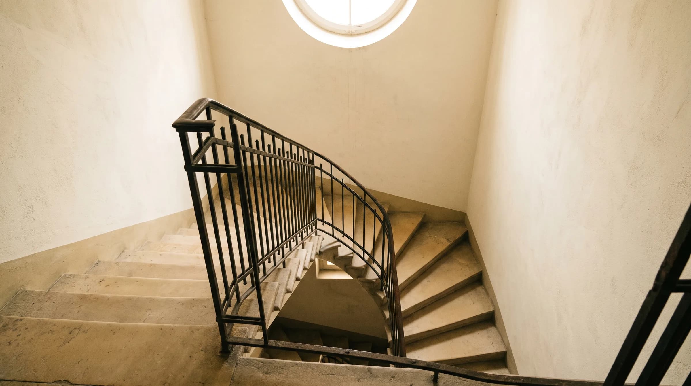 Treppenhaus eines Altbaus mit abgetretenen Sandsteinstufen, schmiedeeisernem Geländer und Tageslicht aus einem Oberlicht.